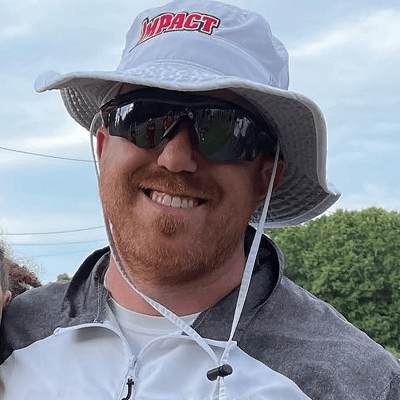 Man wearing sunglasses and a white bucket hat smiles outdoors.