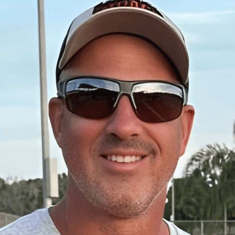 Man wearing sunglasses and a cap smiles outdoors.