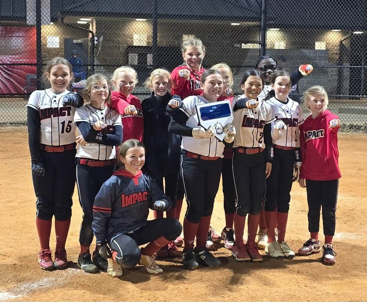 A girls' softball team proudly holding trophies on a field at night.